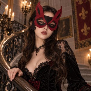 May include: A red and black felt masquerade mask with pointed ear-like extensions, worn with a black velvet long-sleeved dress featuring intricate black lace trim along the neckline and a dark red pendant necklace with a black lace choker base.