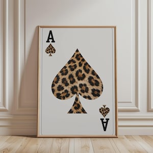 Op de afbeelding: Een ingelijste wandkunstprint met een schoppen aas speelkaartontwerp. Het centrale schoppensymbool en de kleinere schoppenpictogrammen in de hoeken zijn gevuld met een bruin en zwart luipaardpatroon. De letter A is in het zwart gedrukt.