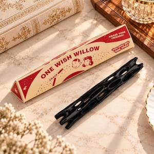 May include: A black, elongated, textured object with multiple oval cutouts, positioned next to a triangular cardboard box. The box is cream and red with the text "ONE WISH WILLOW," "AMAZE YOUR FRIENDS!," and "You only get ONE WISH" printed on it.