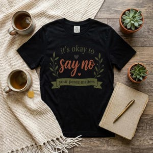 May include: A black short-sleeved t-shirt featuring the text "it's okay to say no" in olive green and "your peace matters" in a dark green banner, accented with small hearts and leafy branches. The shirt is displayed on a wooden surface.