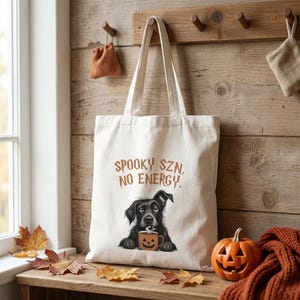May include: A cream-colored canvas tote bag featuring a graphic of a black dog holding a jack-o'-lantern mug. The text above the dog reads SPOOKY SZN, NO ENERGY in a jagged orange font. The bag has two long shoulder straps and a rectangular body.