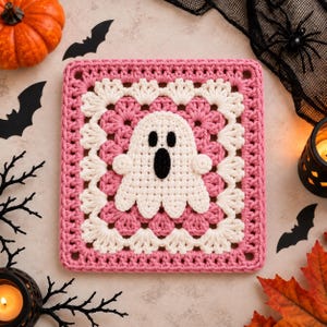 May include: A square crochet granny square featuring a central white ghost applique with black eyes and an oval mouth. The square is bordered by alternating rows of pink and white yarn, with a scalloped white edge surrounding the ghost motif.