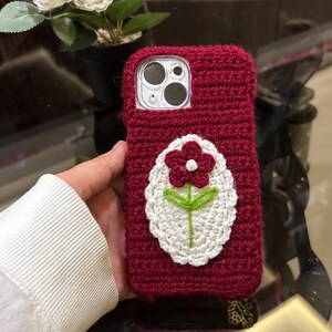 May include: A dark red crochet phone case featuring a central white oval patch with a dark red flower and a green stem. The case has a textured, hand-knitted appearance and includes a cutout for the camera lens on the upper left side.