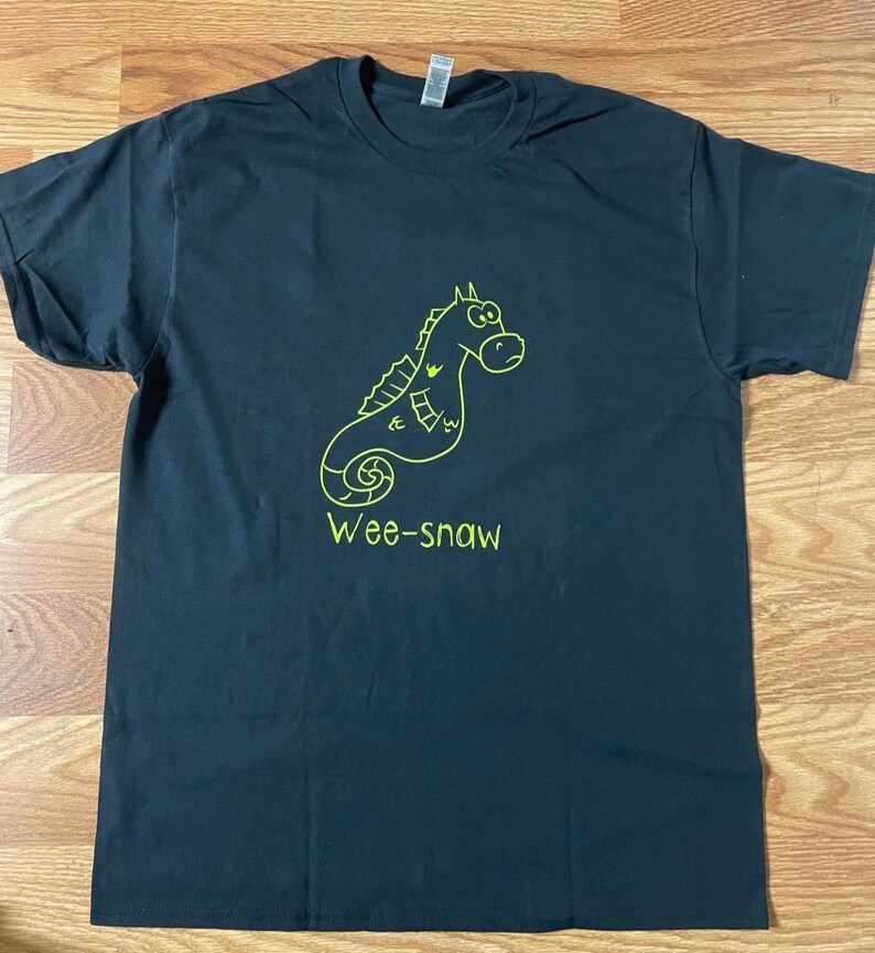 Pode incluir: Camiseta preta com um contorno verde de um cavalo-marinho de desenho animado e o texto "Wee-snaw".