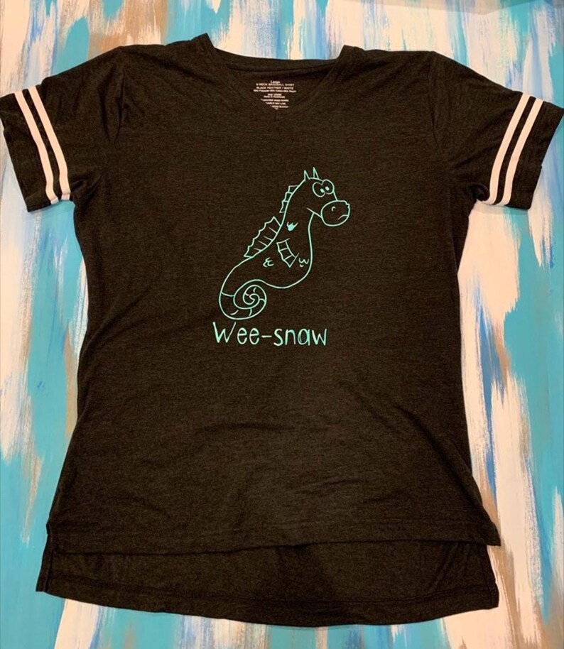 Pode incluir: Uma camiseta cinza escura com um gr&aacute;fico de cavalo-marinho turquesa e o texto "Wee-snaw". A camiseta tem listras brancas nas mangas.