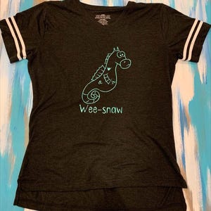 Pode incluir: Uma camiseta cinza escura com um gr&aacute;fico de cavalo-marinho turquesa e o texto "Wee-snaw". A camiseta tem listras brancas nas mangas.
