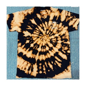 Könnte beinhalten: Schwarzes T-Shirt mit einem Bleich-Tie-Dye-Muster in Braun- und Weißtönen. Das Shirt hat eine kleine Grafik eines Comic-Charakters mit dem Text "GOOD VIBES ONLY" auf der Vorderseite.