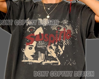 Puede incluir: Una camiseta de manga corta en color gris carbón con un estampado gráfico que incluye el texto rojo Suspiria, una ilustración estilizada de un rostro y elementos arquitectónicos. La prenda tiene cuello redondo y un corte holgado.