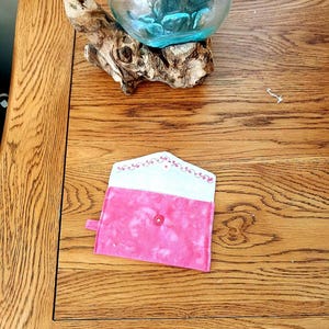 May include: A small, rectangular, pink fabric pouch with a flap closure. The exterior features a mottled pink pattern, while the interior flap is white with a decorative pink floral vine design. A single pink snap button secures the pouch.