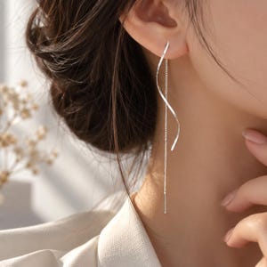 May include: A silver threader earring featuring a long, delicate chain and a smooth, spiralling metal ribbon element. The earring hangs vertically from the earlobe, showcasing a polished, reflective surface and a slender, minimalist design.