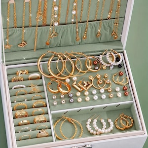 Può includere: Un portagioie bianco con interno verde salvia contenente collane color oro appese a ganci, vari orecchini a cerchio dorati, orecchini con perle, orecchini a lobo e diversi anelli color oro organizzati in file.