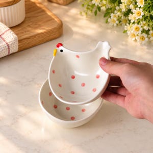 Könnte beinhalten: Eine cremefarbene Keramikschale in Form eines Huhns mit kleinen rosa Punkten, einem roten Kamm und einem gelben Schnabel. Die Schale hat ein glattes, abgerundetes Inneres und eine markante Hühnersilhouette, mit einer passenden Schale darunter.