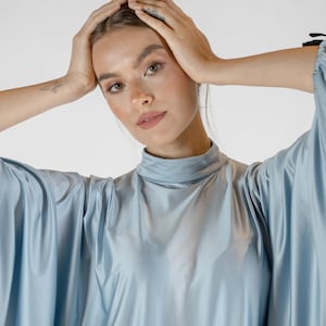 Peut inclure: Un chemisier en satin bleu clair doté d'un col montant et de manches amples et fluides. Le tissu présente une finition lisse et réfléchissante qui capte la lumière, créant des plis et des drapés doux sur le vêtement. Le design est simple et uni.