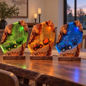 Op de afbeelding: Drie decoratieve lampen van hars en hout, elk met een miniatuurtractor en landbouwapparatuur erin. De lampen gloeien in groen, oranje en blauw, met natuurlijke houten randen en rechthoekige houten voetstukken op een houten tafel.
