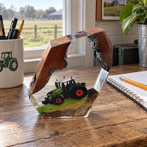 May include: A hexagonal decorative piece made of wood and clear resin, featuring a miniature green tractor with red wheels positioned on a small patch of green grass and brown soil inside the frame, set against a natural wood grain border.