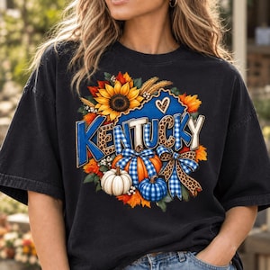 May include: A black short-sleeve t-shirt featuring a graphic print of the word KENTUCKY in blue and leopard print letters. The design includes a large yellow sunflower, orange and blue plaid pumpkins, autumn leaves, and a blue plaid bow.