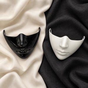 May include: Two half-face masks, one glossy black and one glossy white, each featuring a sculpted nose and lips. The masks are shaped to cover the lower portion of the face and include side loops for attachment, presented as a pair of distinct items.