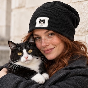 May include: A black waffle-knit beanie featuring a rectangular white patch on the front. The patch displays a graphic illustration of a black and white cat with the text OREO printed in black capital letters underneath the cat image.
