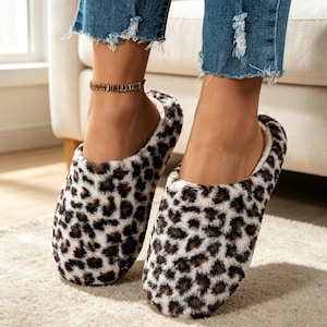 May include: A pair of slip-on slippers featuring a plush, faux-fur texture with a black, brown, and white leopard print pattern. The slippers have a rounded toe design and a soft interior lining, providing a comfortable fit for indoor use.