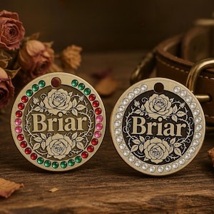 以下が含まれることがあります： セリフ体で「Briar」と刻印され、バラと蔓のエンボス加工が施された2つの円形金属製ペット用IDタグ。左側のタグは赤、ピンク、緑のラインストーンで縁取られ、右側のタグは透明なラインストーンで縁取られています。