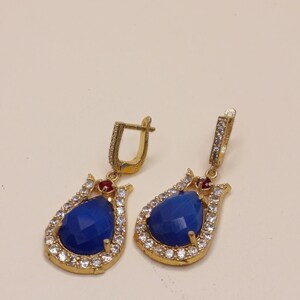 Puede incluir: Un par de pendientes colgantes dorados con una piedra azul central en forma de pera rodeada por un halo de pequeños cristales transparentes. Cada pendiente incluye una pequeña piedra roja en la parte superior y un cierre de bisagra con cristales.