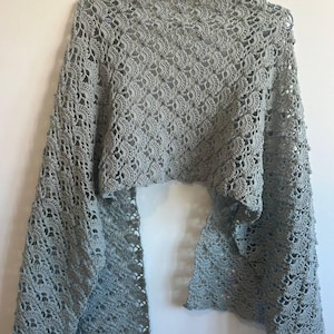 Peut inclure: Un châle au crochet gris clair présentant un motif complexe et répétitif en forme d'éventail sur tout le tissu. Le vêtement est présenté sur un cintre, mettant en valeur son design ajouré, ses bords festonnés et sa structure légère.