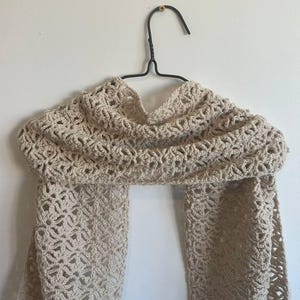 May include: A beige crochet scarf featuring an intricate, repeating openwork pattern. The scarf is draped over a black metal hanger, showcasing the detailed texture of the yarn and the consistent, airy stitch design throughout the entire piece.