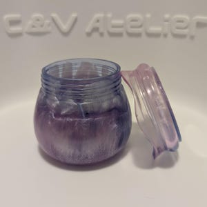 Könnte beinhalten: Ein kleines, rundes, durchscheinendes Gefäß mit violetten und silbernen Wirbelmustern und einem passenden Deckel, der daneben liegt. Das Gefäß hat einen Gewindehals. Im Hintergrund ist der geprägte Schriftzug C&V ATELIER in hellem Ton.