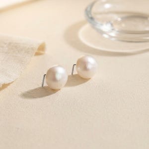 Puede incluir: Un par de pendientes de botón con perlas redondas, blancas y brillantes unidas a postes de metal plateado. Las perlas tienen una superficie lisa y reflectante, colocadas una al lado de la otra para mostrar su forma esférica y brillo natural.