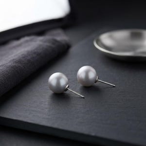 Puede incluir: Un par de pendientes de botón con perlas blancas redondas de acabado liso y brillante. Cada perla está montada de forma segura sobre un fino poste de metal plateado, diseñado para orejas perforadas, mostrando una estética de joyería clásica.