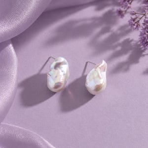 Puede incluir: Un par de pendientes de botón con perlas barrocas blancas de forma irregular y orgánica, con un acabado lustroso e iridiscente. Cada perla cuenta con un poste metálico y presenta una superficie texturizada única con sutiles tonos rosados y azulados.