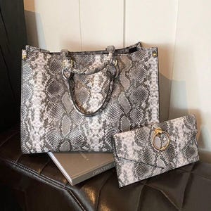 May include: A structured tote bag and a matching envelope clutch, both featuring a grey, black, and white snakeskin pattern. The tote has two top handles with gold-tone hardware, while the clutch includes a prominent gold-tone circular ring.