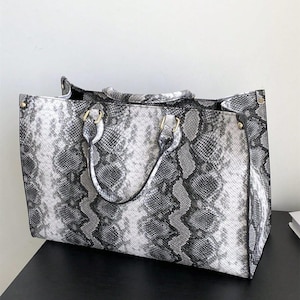 May include: A structured tote bag featuring a grey, black, and white snakeskin pattern. The bag has two top handles attached with gold-toned hardware, a rectangular silhouette, and reinforced corners with small gold-toned rivets on the sides.