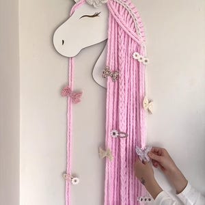 May include: A white unicorn wall hanging featuring a long, flowing mane made of thick pink yarn. The unicorn head has a single horn and closed eyes. The yarn mane serves as a display for various hair clips, including bows and flower-shaped designs.