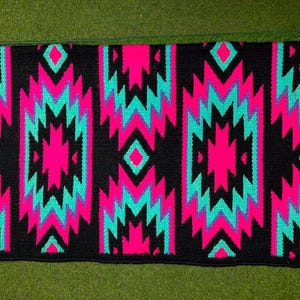 Può includere: Un sottosella rettangolare caratterizzato da un motivo geometrico ripetitivo in rosa neon vibrante e turchese brillante su uno sfondo nero solido. Il design è composto da forme a diamante e chevron impilate disposte in colonne verticali.