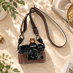 Pode incluir: Uma câmara Fujifilm X-T30 preta dentro de uma capa protetora de couro castanho com costuras brancas. Uma pequena figura de urso castanho está fixada no topo da câmara. O dispositivo possui uma correia de couro castanho longa e ajustável.