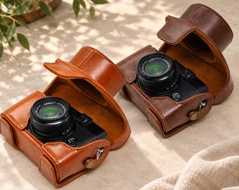 Könnte beinhalten: Zwei schwarze Digitalkameras befinden sich jeweils in einer schützenden braunen Ledertasche. Die Taschen haben ein strukturiertes Design mit einer oberen Klappe, die sich zurückklappen lässt und das Objektiv sowie das Gehäuse freigibt. Metall-Druckknopf.
