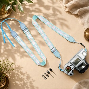 May include: A light blue camera strap featuring a marbled, iridescent pattern in pastel tones. The strap includes silver-toned metal hardware, adjustable sliders, and leather-like end tabs. A silver camera body is attached to one end of the strap.