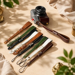 Pode incluir: Uma câmara digital prateada com uma bolsa de couro castanho presa a uma correia de pulso de couro castanho, juntamente com uma coleção de oito correias adicionais em várias cores, incluindo castanho, verde, preto e branco, com costuras.