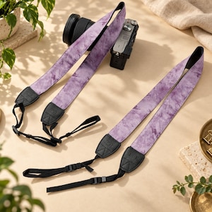 Könnte beinhalten: Zwei Kameragurte mit violettem Batikmuster und schwarzen kunstlederartigen Endstücken. Ein Gurt ist an einer schwarzen Digitalkamera befestigt, der andere liegt flach. Die Endstücke tragen die eingeprägten Schriftzüge Taylor und J K.