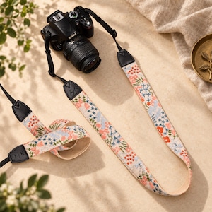 May include: A black Nikon DSLR camera attached to a long, fabric camera strap featuring a colourful floral embroidery pattern on a cream background. The strap includes black leather end tabs, one embossed with the name Taylor and another with J K.