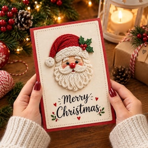 Puede incluir: Una tarjeta de felicitación rectangular con frente color crema que presenta un rostro de Santa Claus bordado en tres dimensiones con gorro rojo, barba blanca y una ramita de acebo. El texto Merry Christmas está impreso en negro.