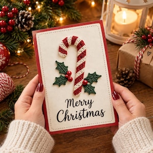 May include: A rectangular greeting card with a red border and a cream-colored center featuring a crocheted candy cane and green holly leaves with red berries. The text Merry Christmas is printed in black script below the candy cane design.