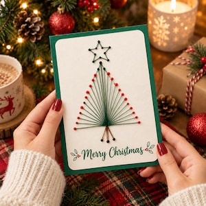 May include: A rectangular greeting card with a dark green border and a white center featuring a string art Christmas tree design. The tree is made of green thread and red beads, topped with a star. The text Merry Christmas is printed in green.