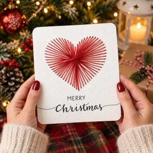 May include: A white rectangular greeting card featuring a red string art heart design in the center. Below the heart, the text reads MERRY Christmas in black lettering. The card has rounded corners and is held against a festive holiday background.