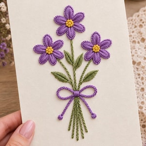 Op de afbeelding: Een rechthoekige gebroken witte kaart met een geborduurd ontwerp van drie paarse bloemen met gele harten. De bloemen hebben groene stengels en bladeren en zijn aan de onderkant van de stengels samengebonden met een paarse strik.
