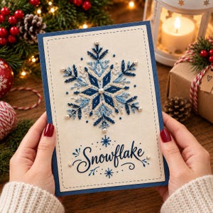 May include: A rectangular greeting card with a dark blue border and a cream-colored center panel. The card features a central embroidered snowflake design in shades of blue and white, accented with small white pearls and the word Snowflake in blue.