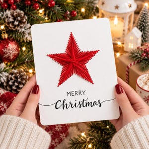May include: A white rectangular greeting card featuring a red star created with string art. The text Merry Christmas is printed in black below the star. The card is held against a background of pine branches, red ornaments, and lights.