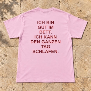 Könnte beinhalten: Ein hellrosa T-Shirt mit kurzen Ärmeln und dunkelrotem Text auf der Rückseite, der lautet: ICH BIN GUT IM BETT. ICH KANN DEN GANZEN TAG SCHLAFEN. Das Shirt hat einen Rundhalsausschnitt, eine Standardpassform und einen geraden Saum.