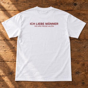 Könnte beinhalten: Ein weißes T-Shirt mit kurzen Ärmeln und dunkelrotem Text auf der Rückseite. Der Text lautet in größerer Schrift ICH LIEBE MÄNNER, gefolgt von dem kleineren Text DIE IHRE FRESSE HALTEN. Das T-Shirt hat einen Rundhalsausschnitt und eine Standardpassform.